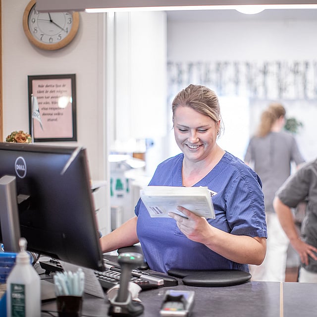 Kvinnlig veterinär står framför en dator och kollar ner mot en anteckningsblock och ler. Bakom henne syns en till glad kvinnlig veterinär och bakom dem finns det ännu en veterinär som står med ryggen mot alla.