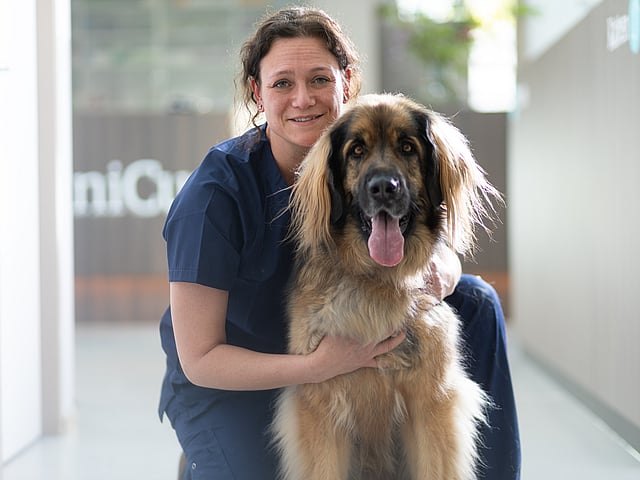 Kvinnlig brunhårig veterinär med blåa ögon sitter på golvet och håller om en långhårig, ljusbrun hund bakifrån.