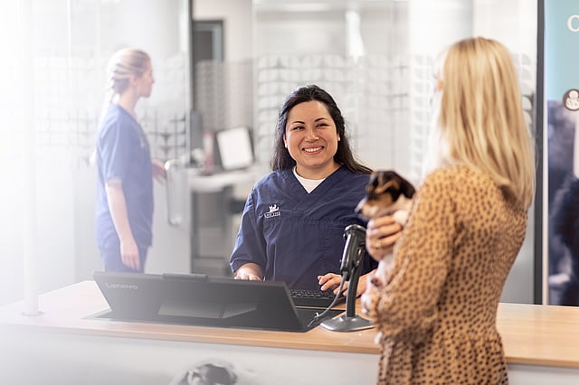 Kvinnlig hundägare håller om sin hund och pratar med en receptionist hos AniCura.